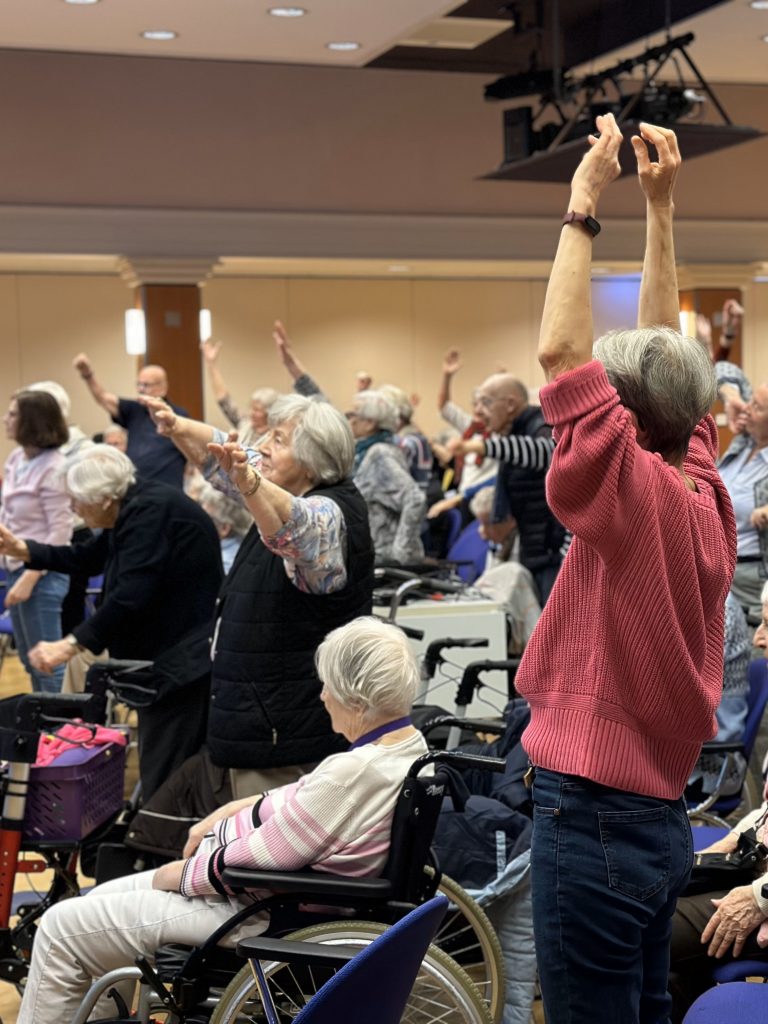 Eine Gruppe von Senior*innen wärmt sich auf. Sie heben die Arme, jede Person so hoch, wie sie kann. 