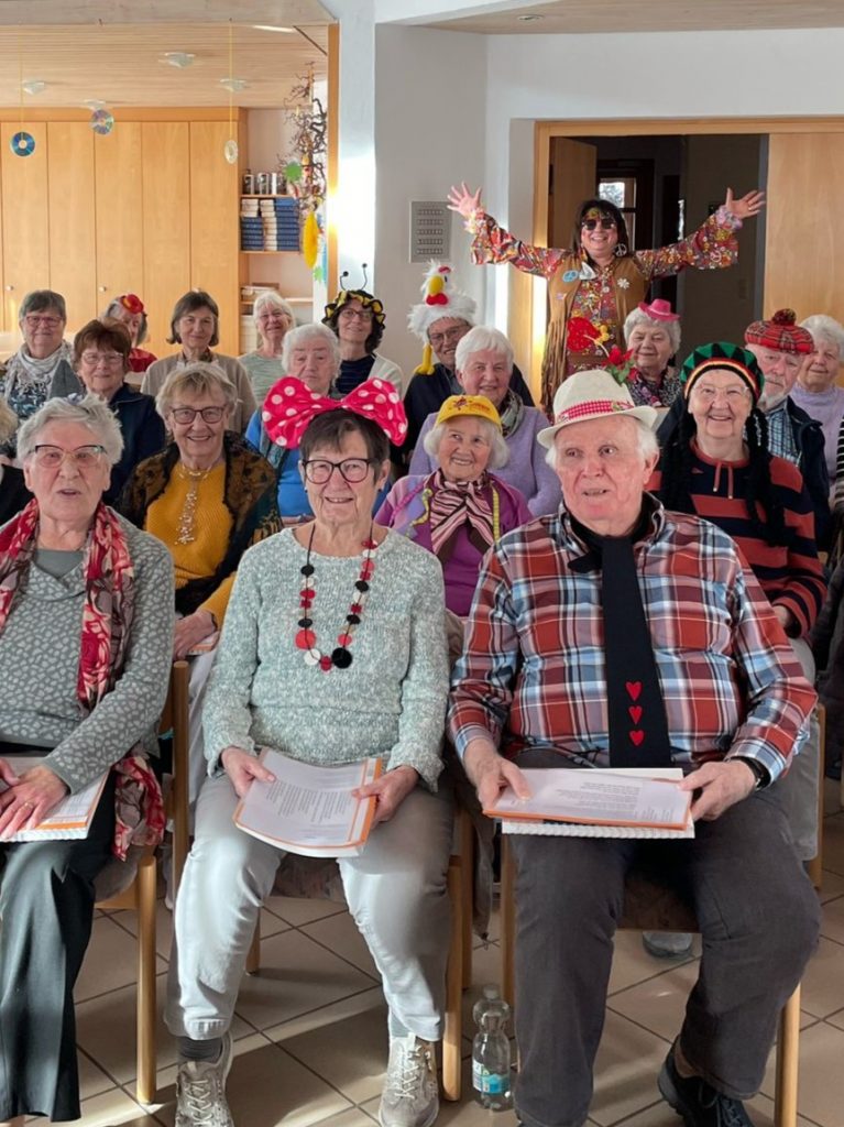 Eine Gruppe Senior*innen sitzt auf Stühlen. Sie sind alle für den Karneval verkleidet und haben Notenbücher in den Händen. Alle lachen in Kamera.