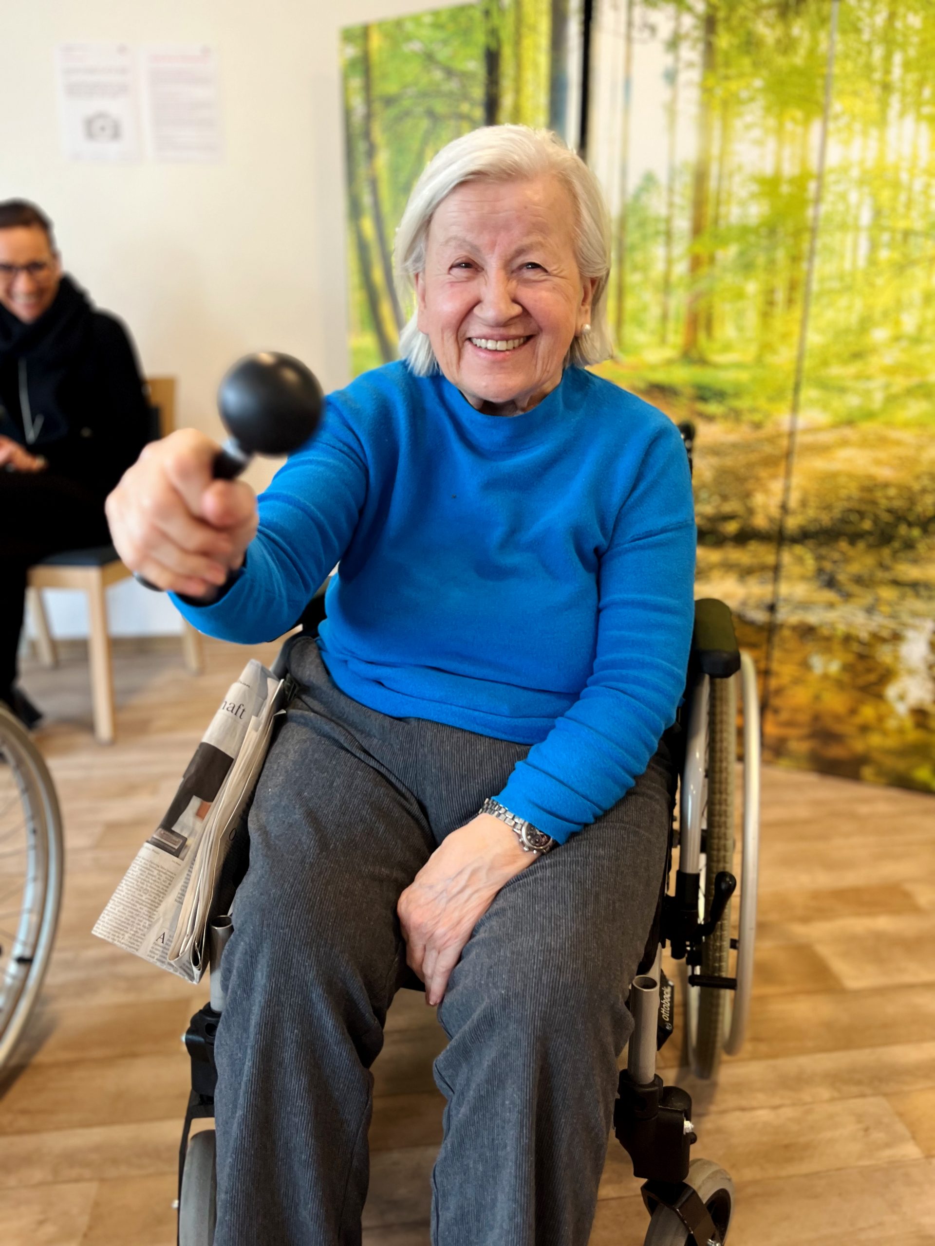 Eine ältere lächelnde Frau im Rollstuhl zeigt ein Rhythmusinstrument.