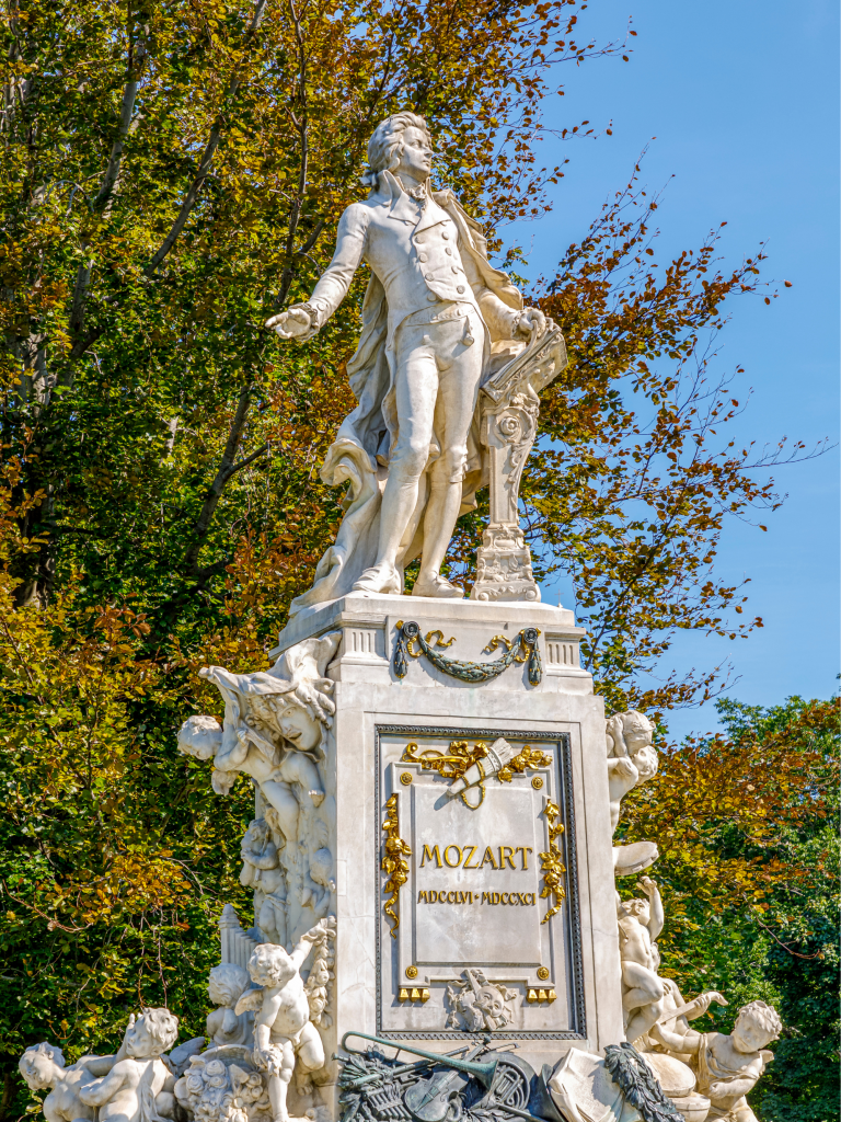 Mozart-Denkmal in Wien: Der Komponist ist mit einem Notenpult auf einem geschmückten Sockel dargestellt. Dieser zeigt Ornamente, Masken, Kränze und Puttenfiguren.