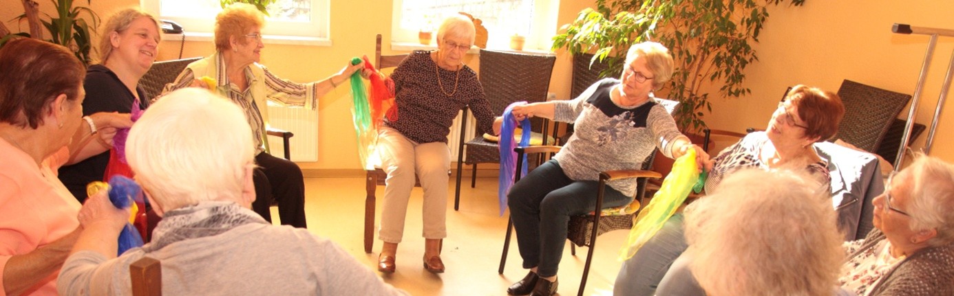 Seniorinnen sitzen im Kreis, halten sich an den Händen und neigen sich gemeinsam nach links. 