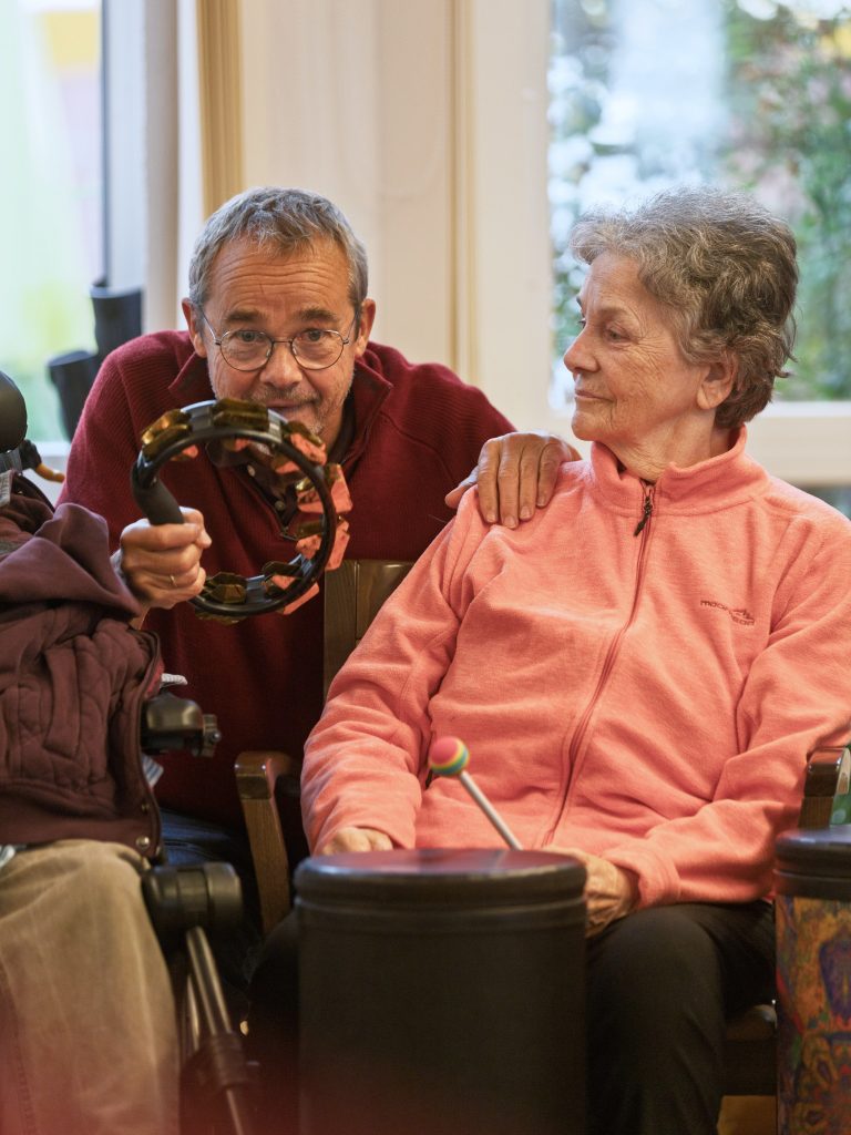 Zwei Senior*innen an Trommel und Schellenkranz. Sie interagieren mit Blicken und Berührung miteinander. 