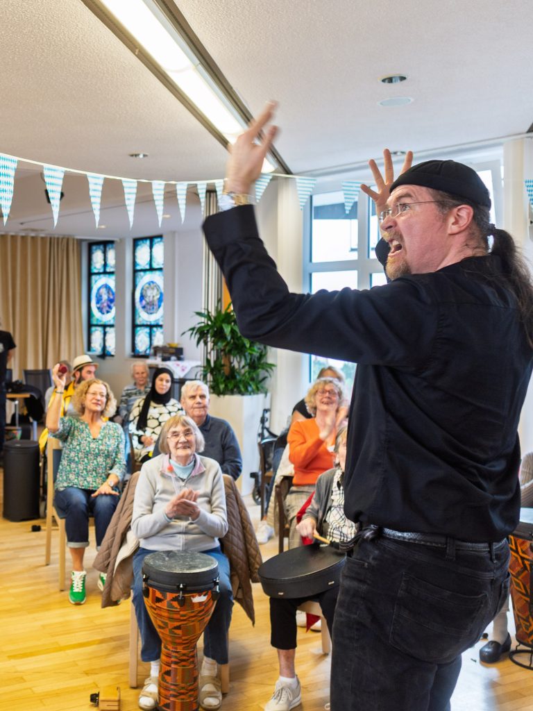 Ein Projektleiter steht vor einer Gruppe lachender und sich bewegender Senior*innen. Er macht ausladene Bewegungen und zieht Grimassen. 
