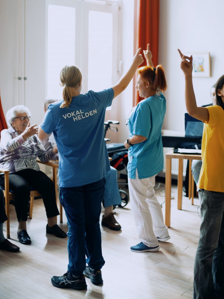 Eine Pflegekraft leitet Bewegungen zum Gesang an. Sie wird unterstützt von einer Projektmitarbeitenden.