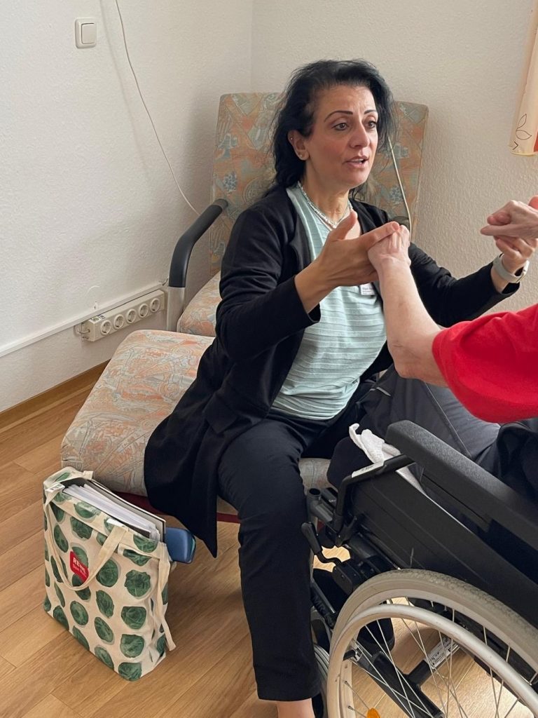 Foto der Projektleitung, die sich auf einen Sessel gesetzt hat, um mit einer Seniorin im Rollstuhl auf Augenhöhe zu sein. Sie fassen sich an den Händen und führen gemeinsam eine Bewegung aus.