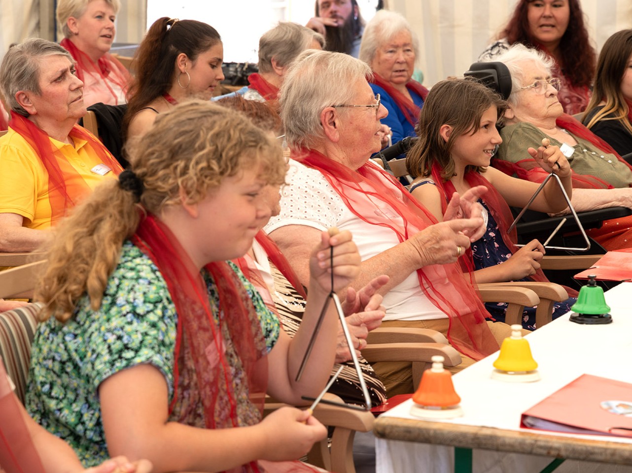 Jugendliche und Senior*innen sitzen bunt gemischt in Reihen. Die Jugendlichen spielen Triangel, die Erwachsenen singen. 