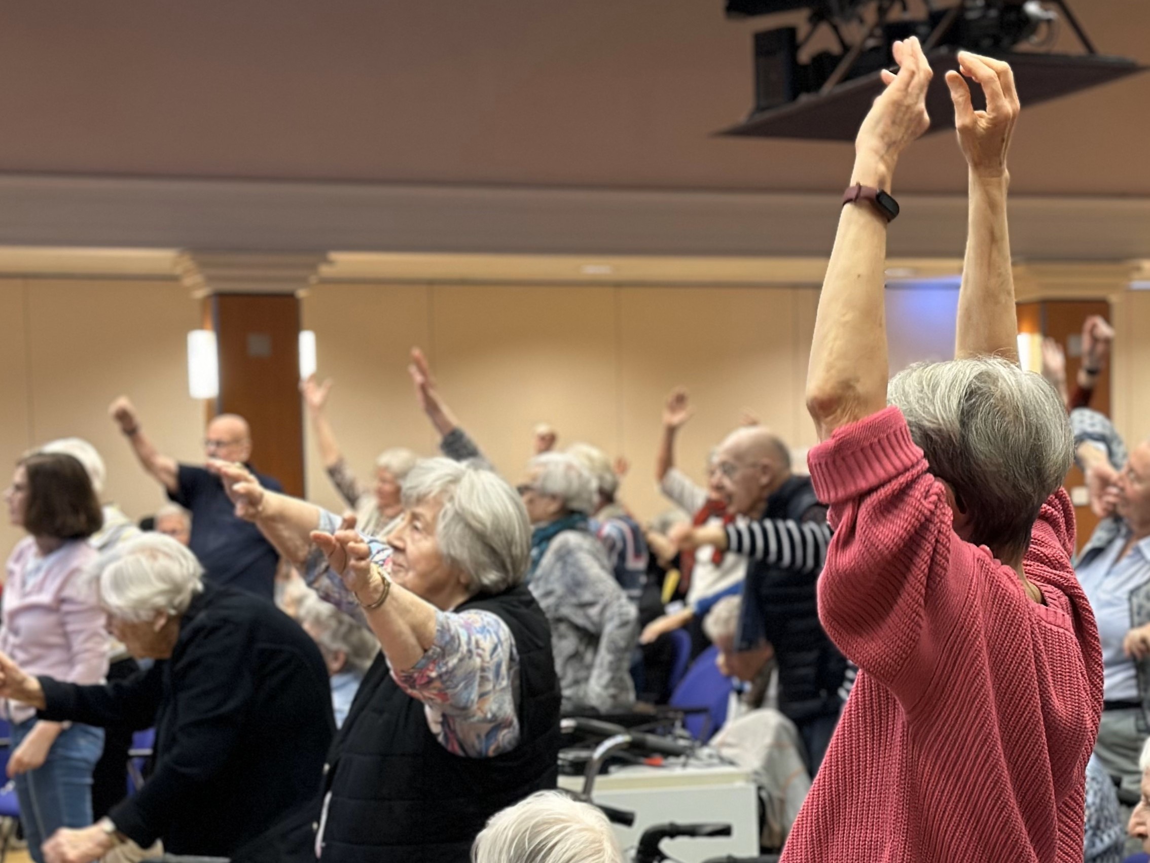 Eine Gruppe von Senior*innen wärmt sich auf. Sie heben die Arme, jede Person so hoch, wie sie kann.