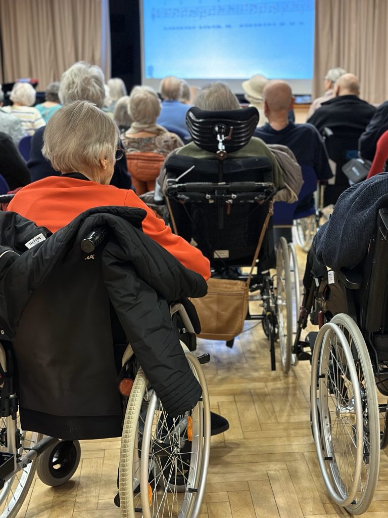 Eine Gruppe von Senior*innen (teils im Rollstuhl) sitzen vor einer Leinwand mit Liedtext.