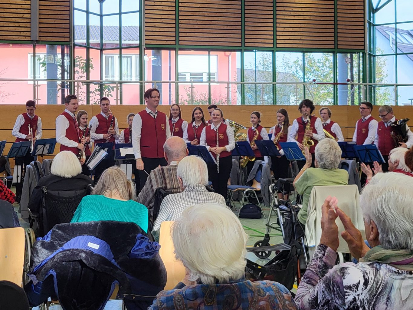 Eine Turnhalle. Das Blasmusikensemble trägt Uniform und hat sich erhoben. Das Publikum: Senior*innen, die klatschen.