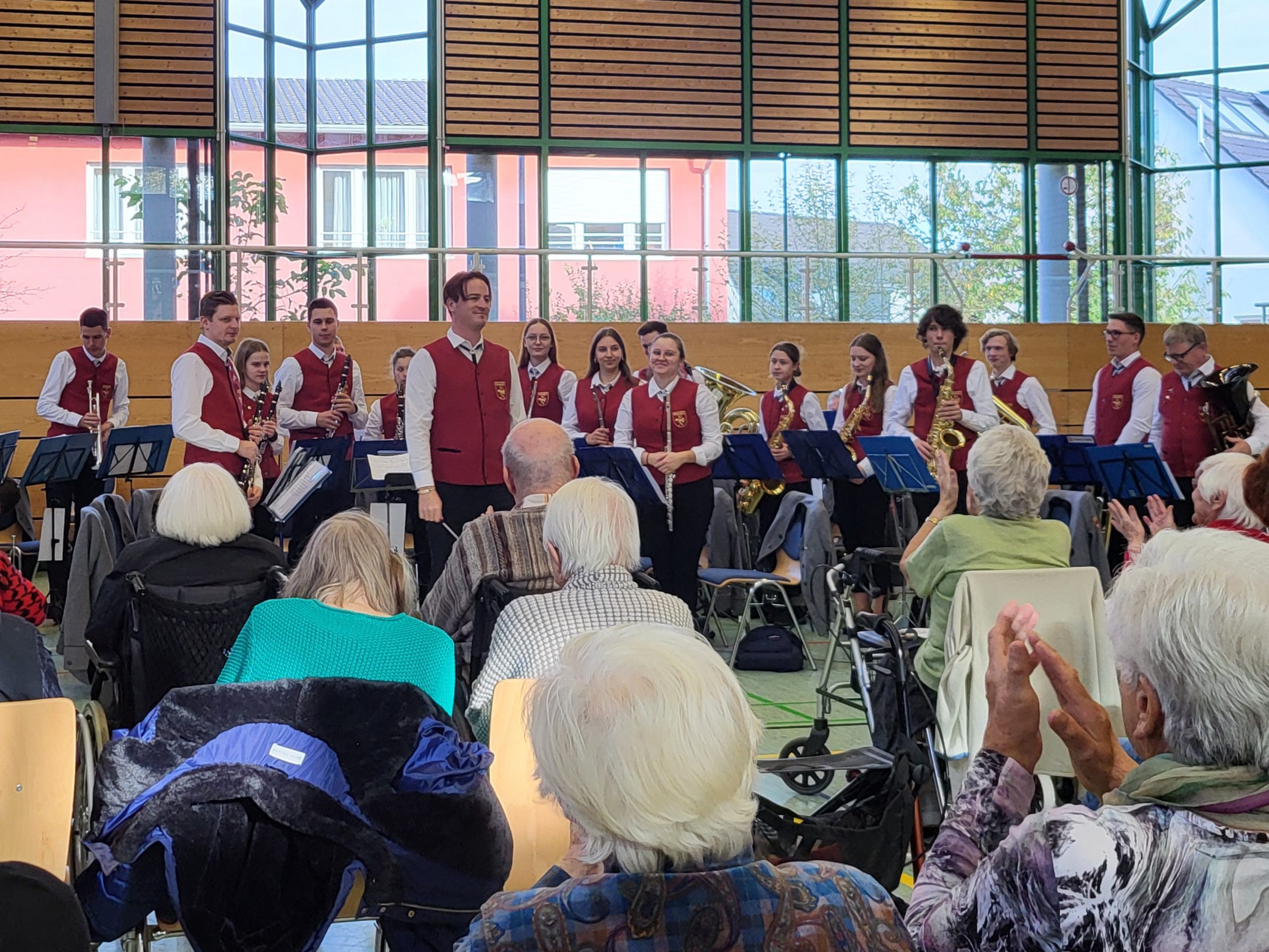Ein Musikensemble in Uniform spielt in einer Turnhalle. Das Publikum: Senior*innen, die klatschen.