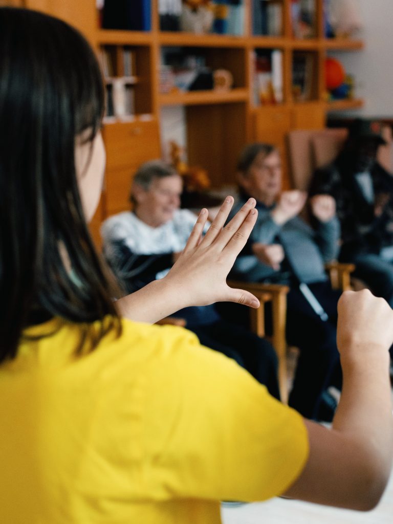 EIn Kind zeigt mit den Händen Tanzbewegungen vor. Senior*innen ahmen sie nach. 