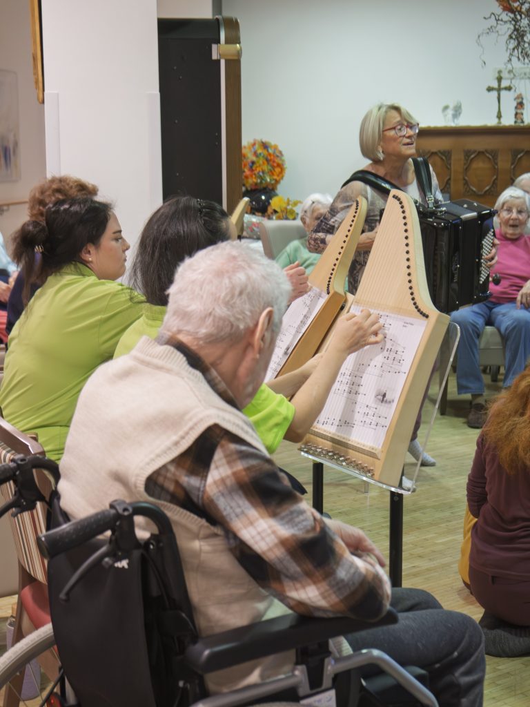 Senior*innen sitzen singend im Kreis, Pflegepersonal spielt Tischharfen, eine Akkordeonspielerin steht in der Mitte.