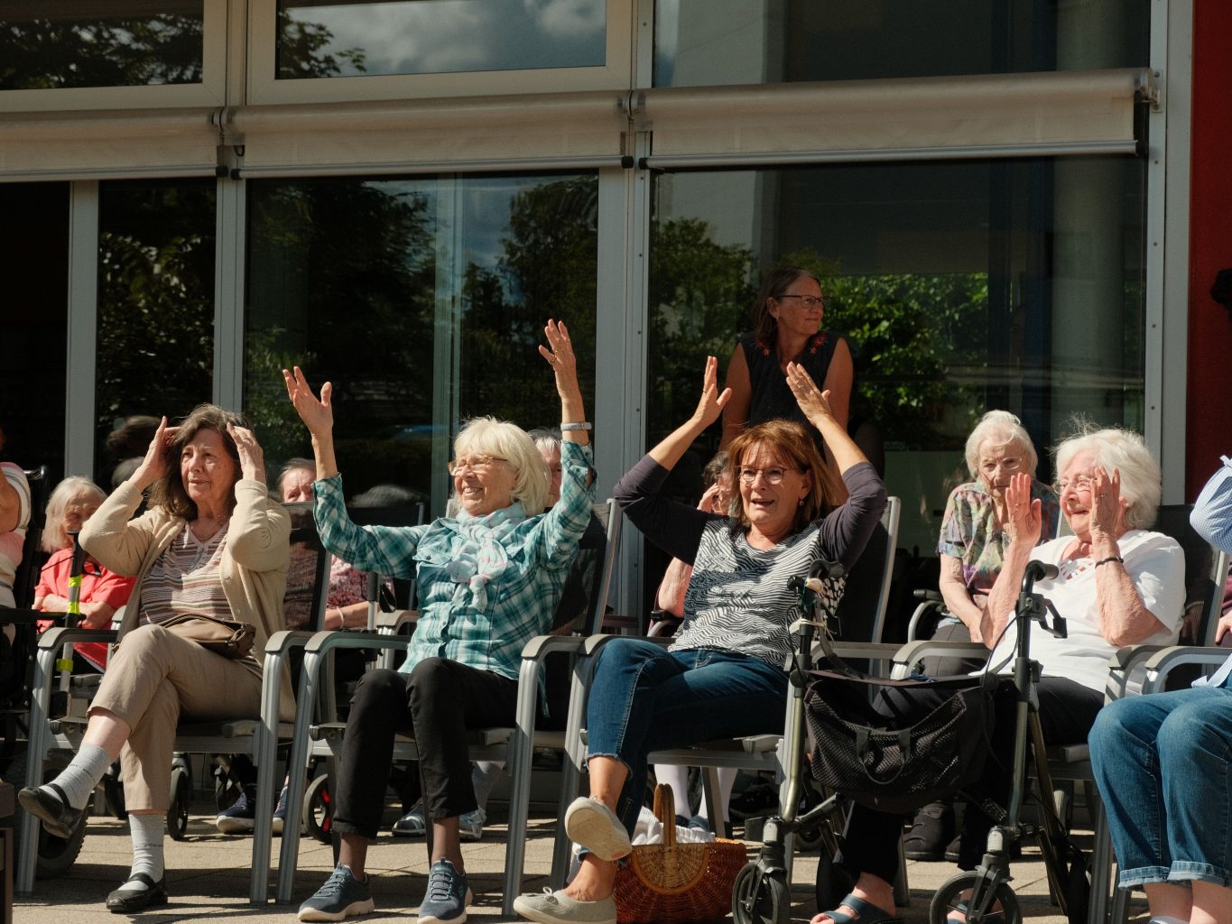 Seniorinnen machen gemeinsam Bewegungen im Sitztanz. Sie sitzen draßen, die Sonne scheint.