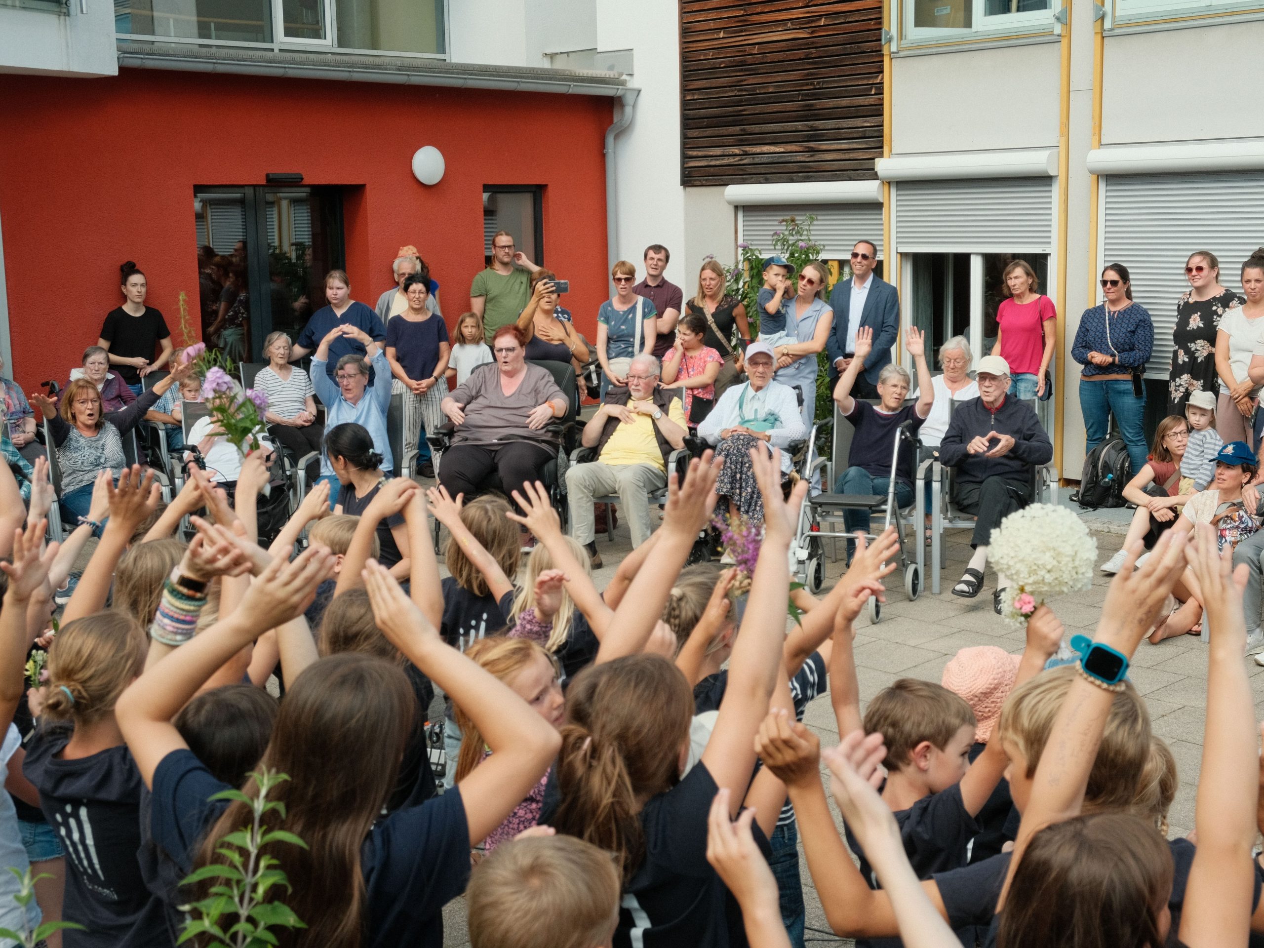 Eine Gruppe Kinder und eine Gruppe Senior*innen stehen sich gegenüber und führen zusammen Tanzbewegungen mit den Händen aus.