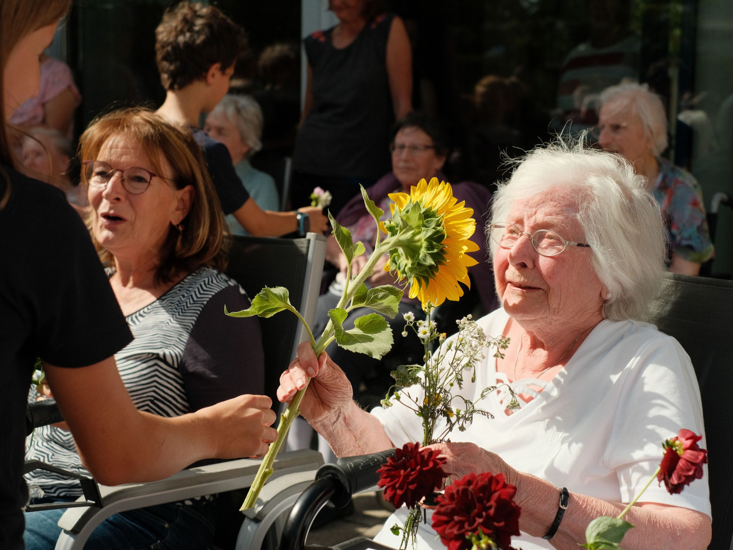 Ein Kind gibt einer Seniorin eine Sonnenbume in die Hand, die Seniorin lächelt berührt.