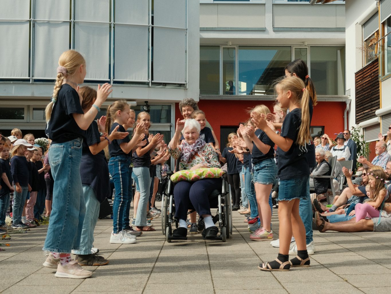 Kidner bilden ein Spalier. Eine Seniorin im Rollstuhl wird von einer Betreuerin und einem Kind durch das  Spalier gefahren. Die Kinder klatschen, die Seniorin lacht herzlich. 