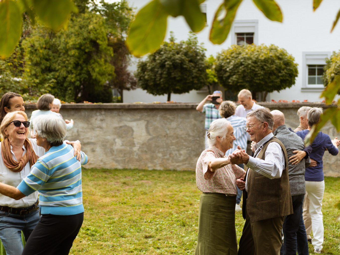 Paare von unterschiedlich alten Menschen tanzen auf einer Wiese. Sie lachen. 