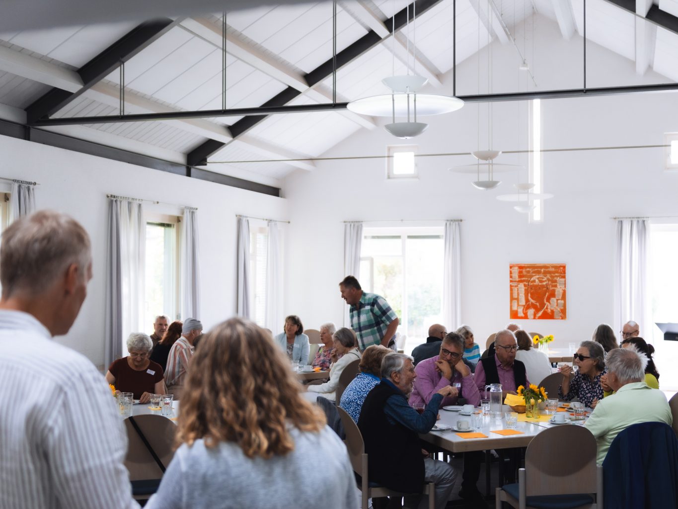 Blick in das Café: Ein moderner. heller Raum. Menschen sitzen an runden Tischen mit Sonnenblumen, unterhalten sich.