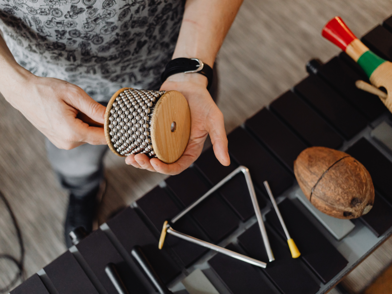 Verschiedene Rhythmusintrumente liegen nebeneinander: Triangel, Rassel, Guiro. Eine Hand hält eine Cabasa.