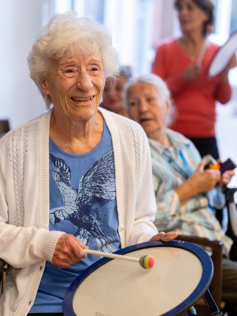 EIne ältere Frau blickt begeistert und lahct, während sie mit einem Schlägel trommelt.