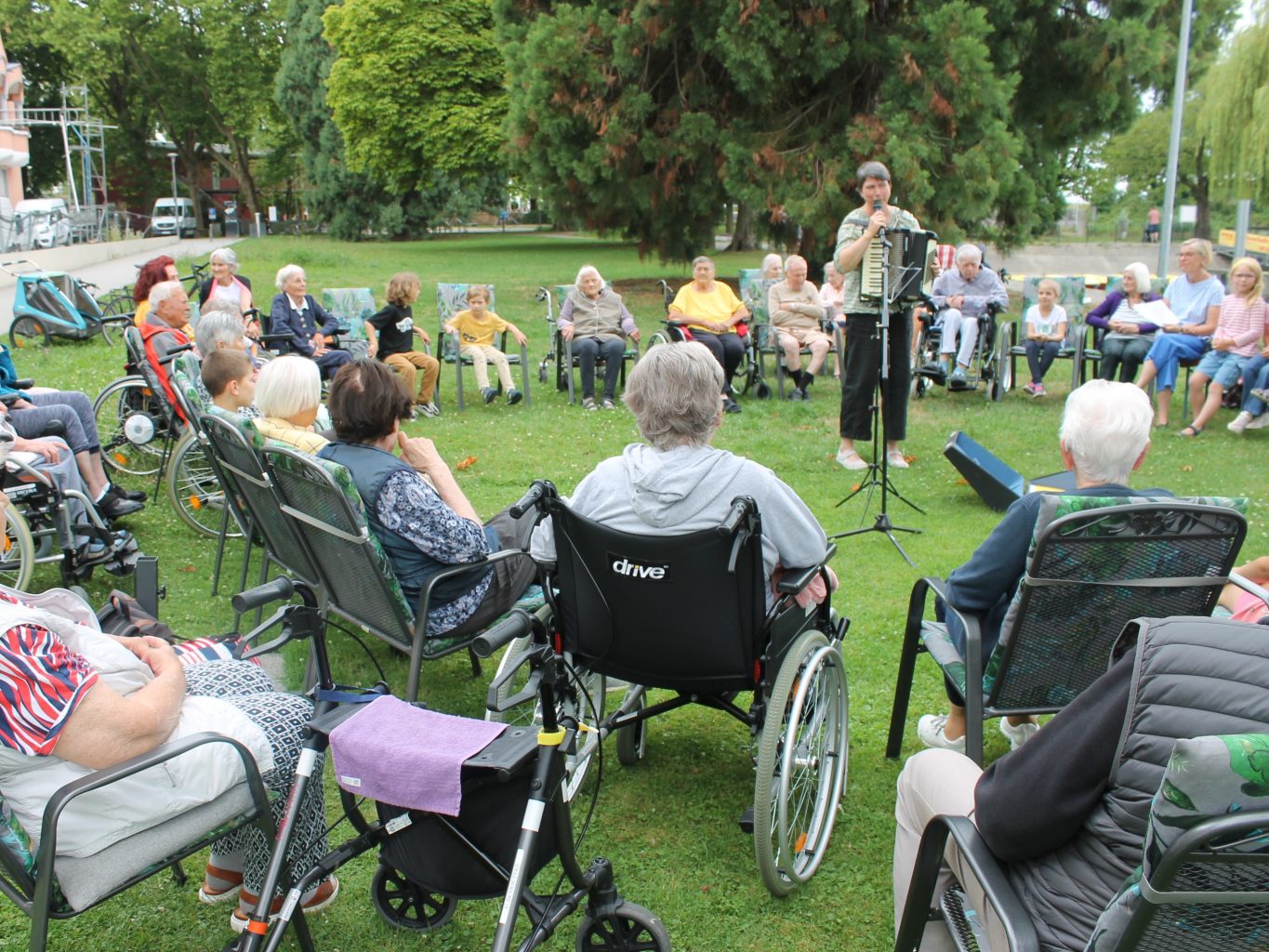 Auf einer grünen Wiese sitzen Senior*innen und Kinder gemischt im Kreis. In der Mitte steht eine Projektmitarbeitende mit Akkordeon. 