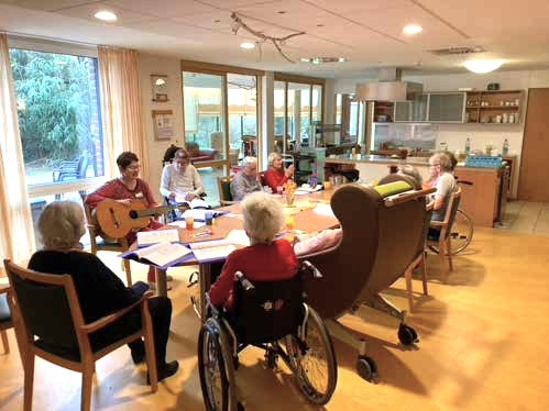 Senior*innen sitzen um einen großen runden Tisch. Eine Projektmitarbeiterin spielt Gitarre. 