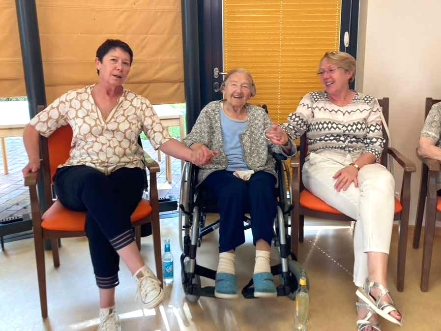 Frauen sitzen im Singkreis, halten sich an den Händen und lachen in die Kamera. 