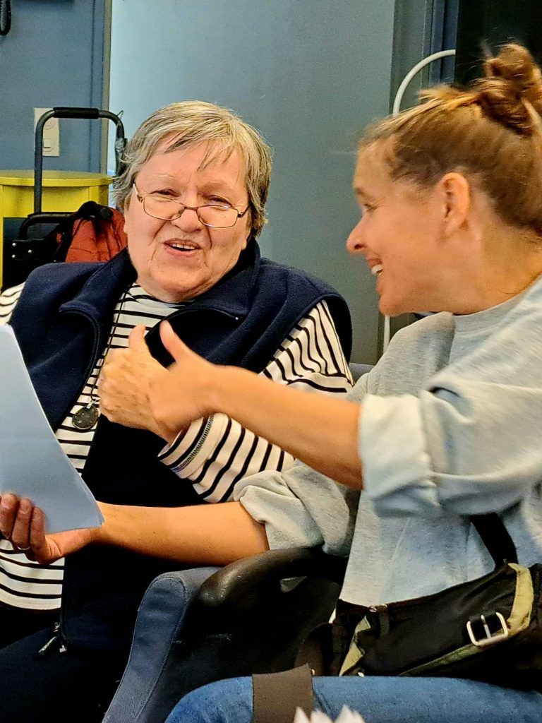 Eine Seniorin und eine Betreuerin schauen in ein Liedblatt, lachen und halten die Daumen hoch. 