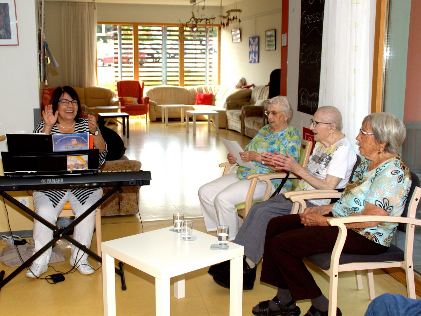 Eine Projektmitarbeiterin sietzt am Keyboard und klatscht. Neben ihr drei Seniorinnen, die engagiert singen und klatschen. 