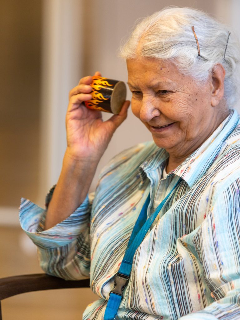 Eine Seniorin hält sich eine kleine Rassel ans Ohr und lächelt.