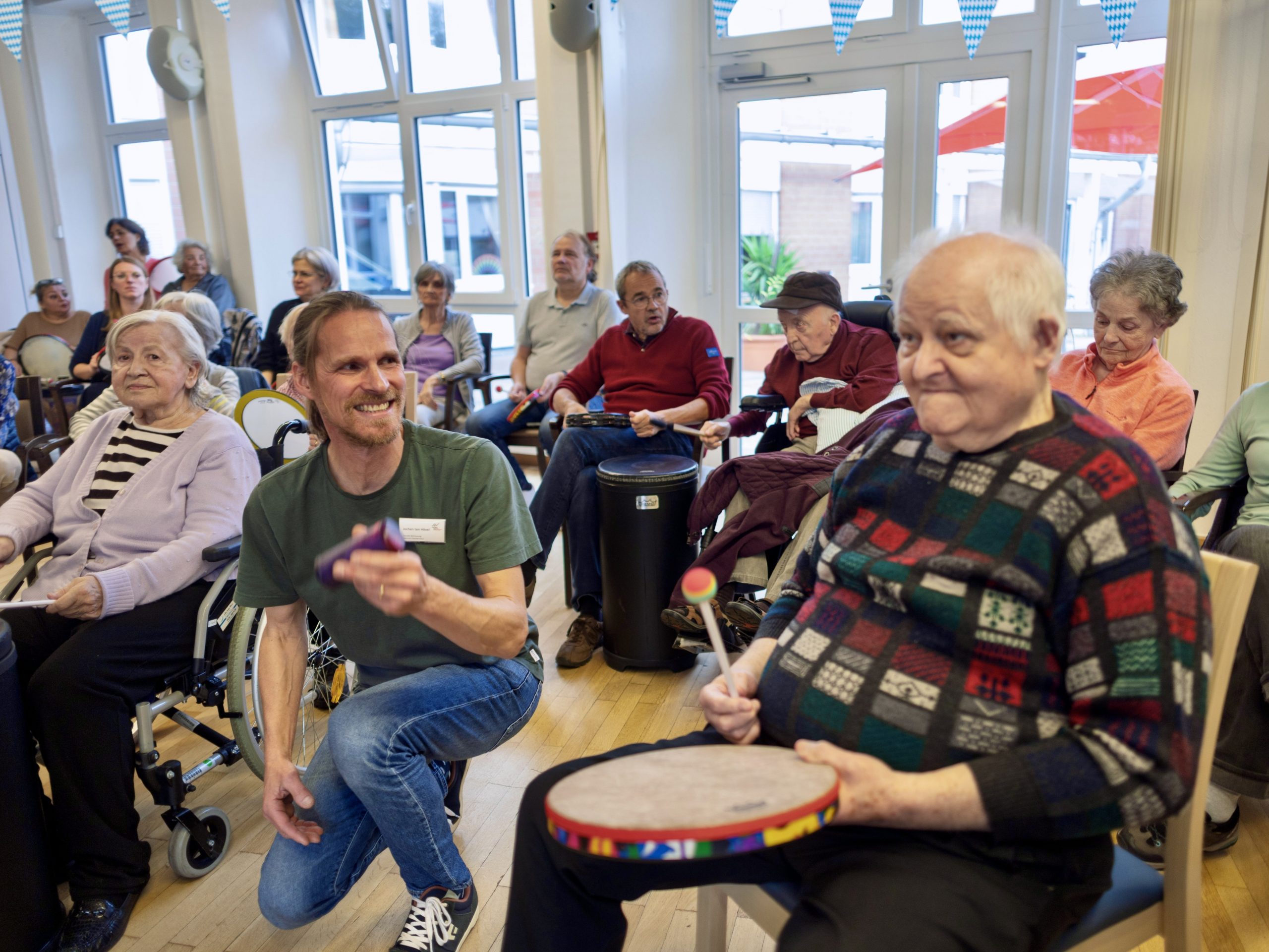 Eine Gruppe Senior*innen trommelt gemeinsam. Der Projektleiter kniet am Boden und grinst in Richung eines Seniors.