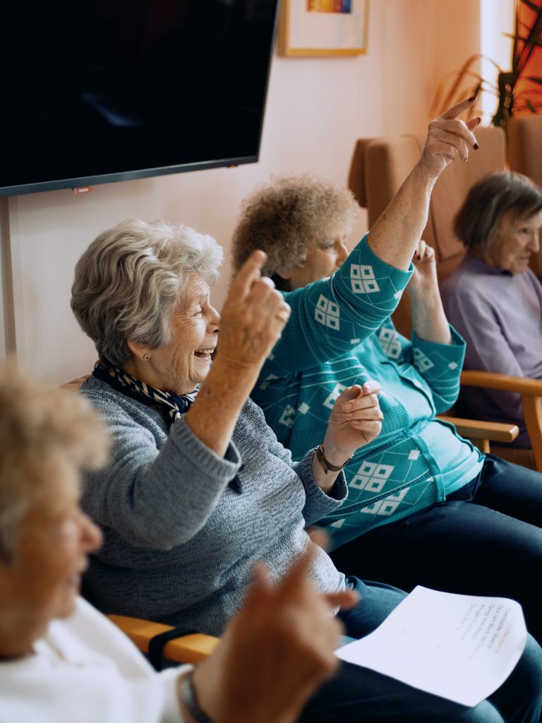 Seniorinnen sitzen in einer Reihe und führen gleichzeitig Bewegungen aus. Sie lachen. 