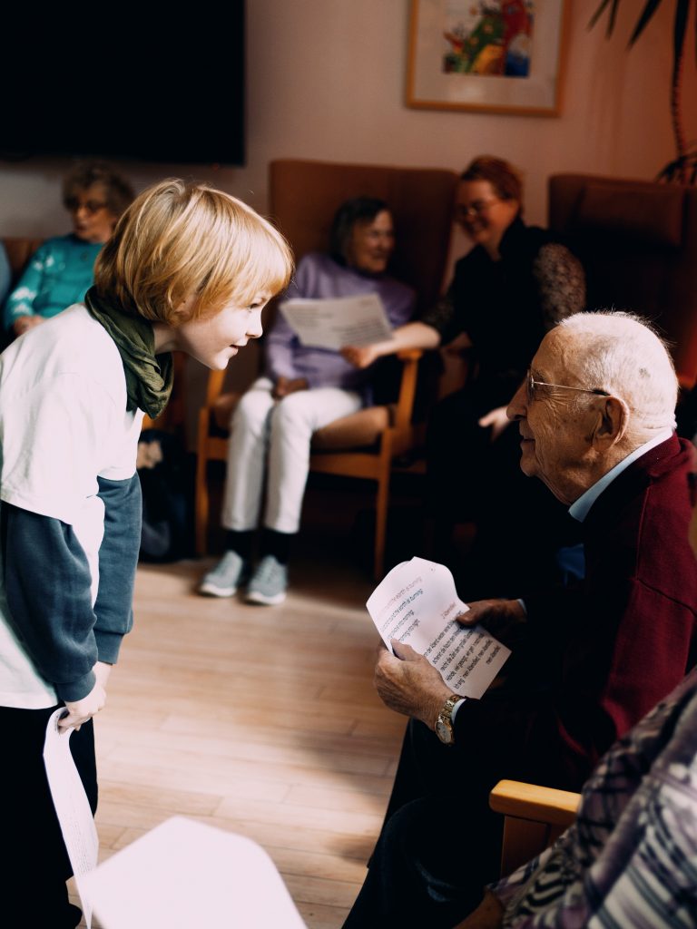 Ein Kind beugt sich zu einem Senior, der mit Notenblatt auf seinem Platz sitzt und das Kind anspricht.