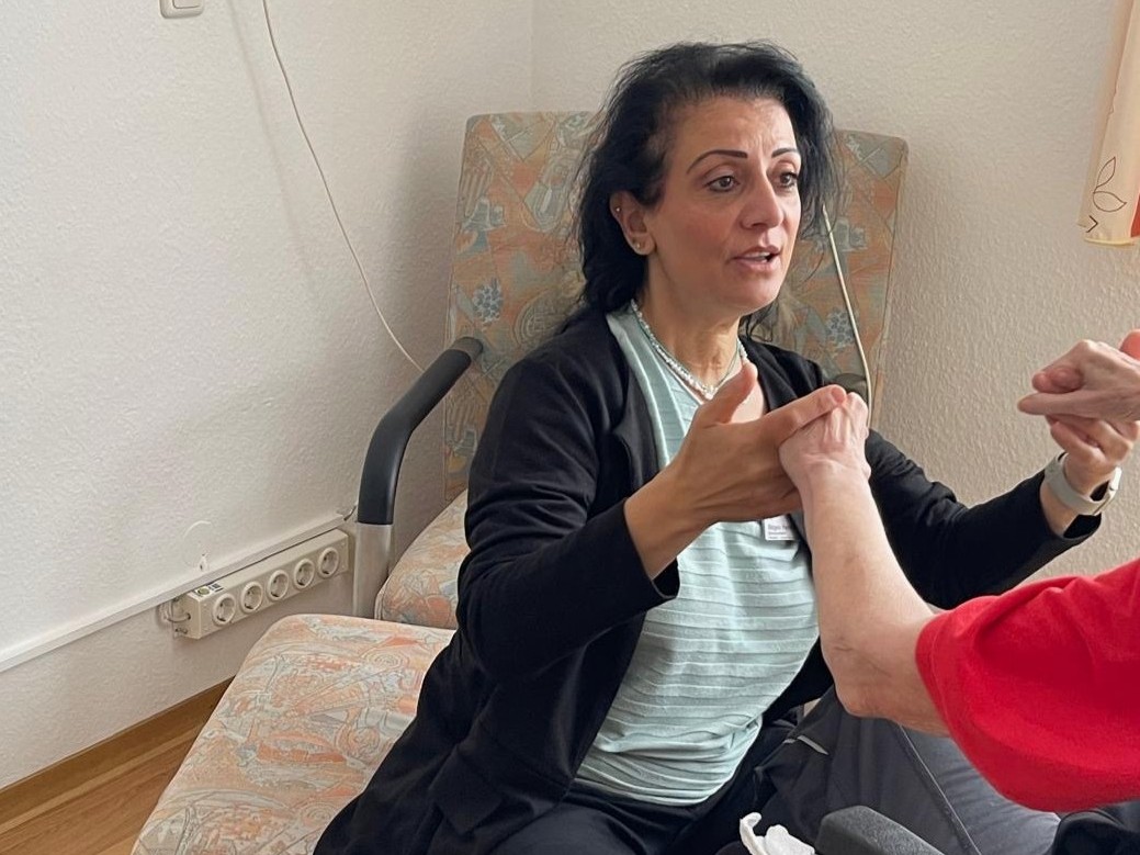 Foto der Projektleitung, die sich auf einen Sessel gesetzt hat, um mit einer Seniorin im Rollstuhl auf Augenhöhe zu sein. Sie fassen sich an den Händen und führen gemeinsam eine Bewegung aus.